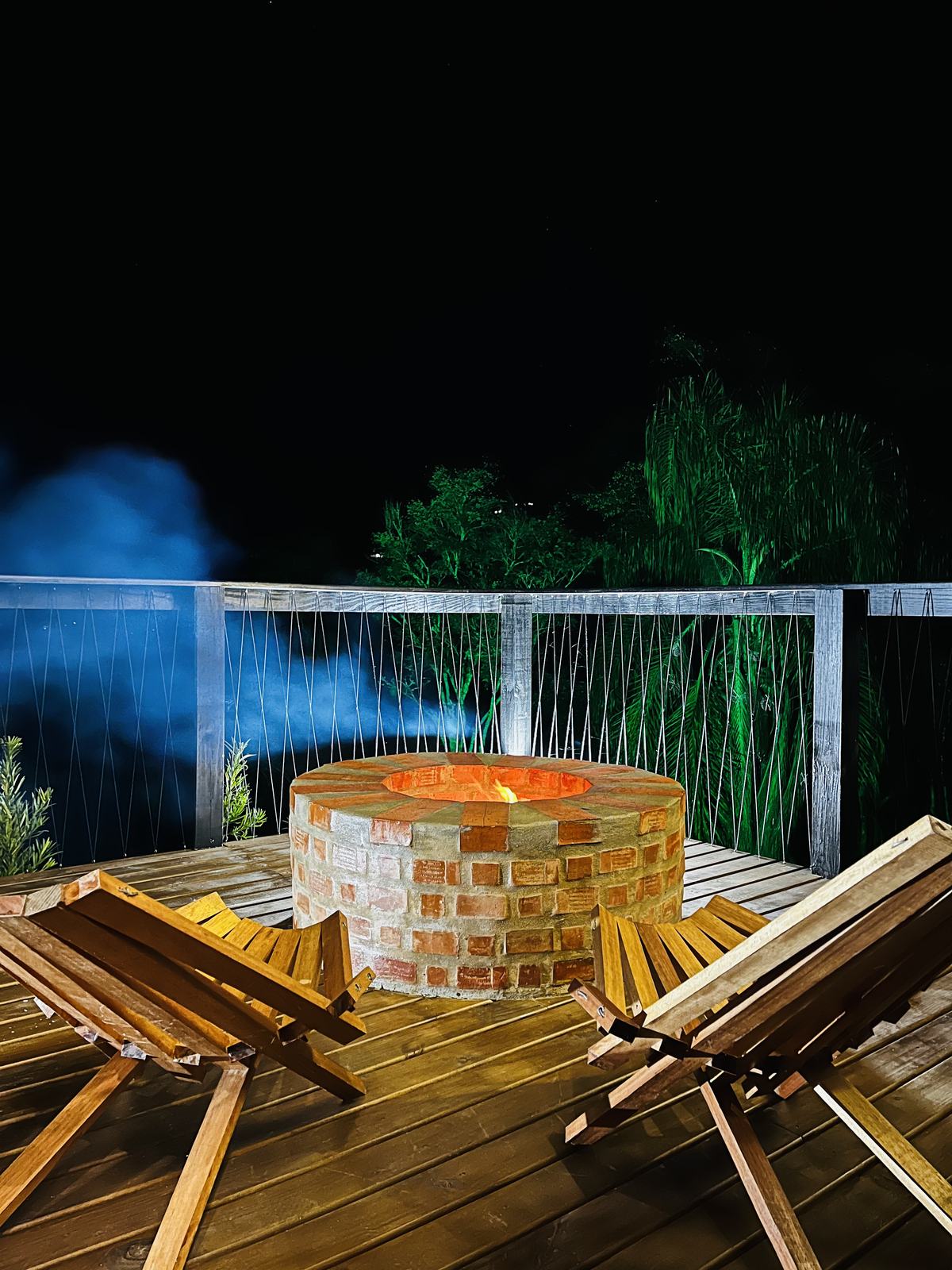 Fogueira acesa à noite entre cadeiras de deck, com fumaça e árvores iluminadas